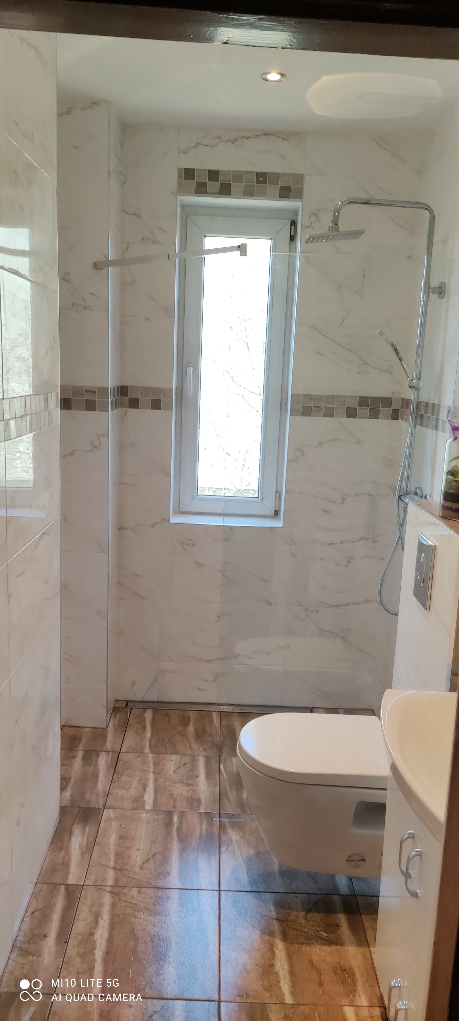 Wykończona łazienka z marmurowymi płytkami na ścianach, brązowymi płytkami podłogowymi i kabiną prysznicową typu walk-in z deszczownicą, widok na okno z dekoracyjnym paskiem mozaiki.