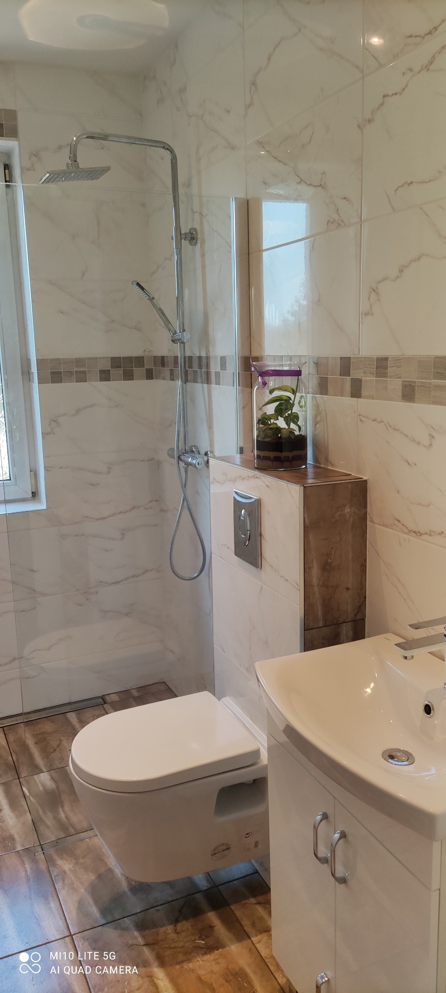 Nowoczesna łazienka z marmurowymi płytkami, wiszącą toaletą, kabiną prysznicową typu walk-in i umywalką z szafką, ozdobiona terrarium z roślinami na zabudowie stelaża.