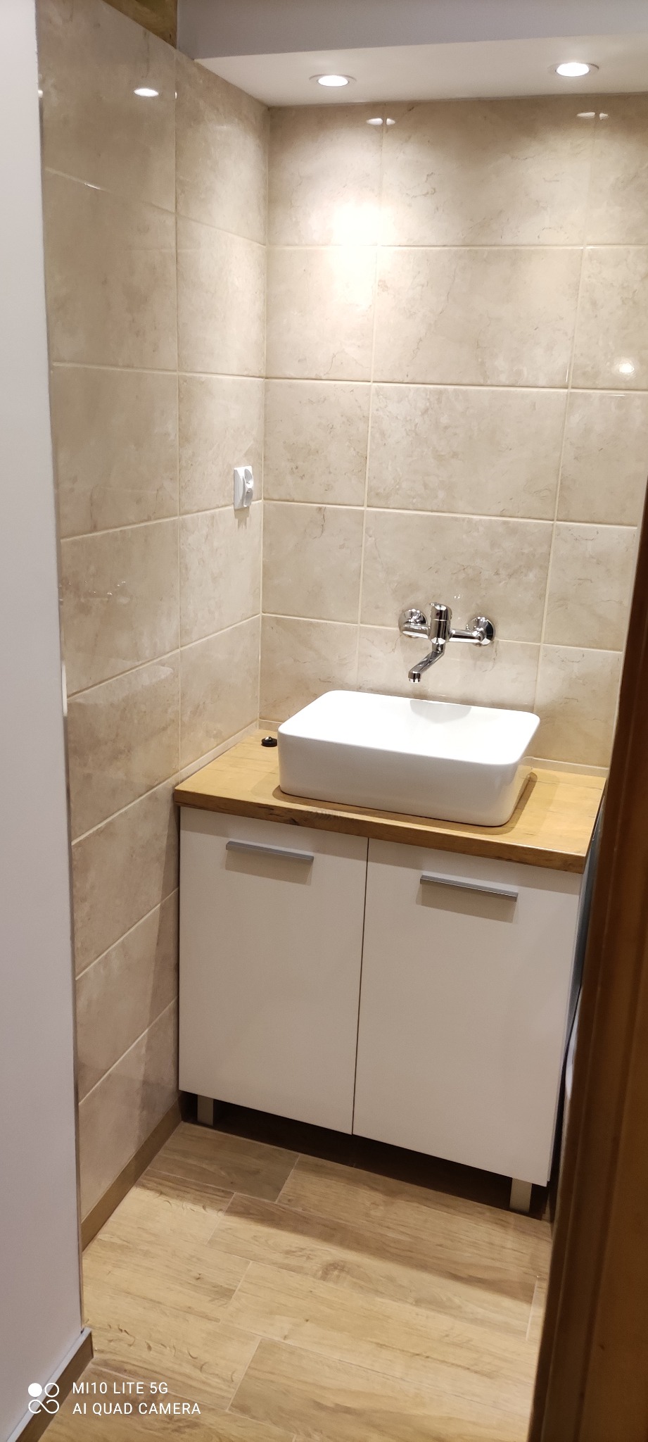 Umywalka nablatowa o kwadratowym kształcie, osadzona na drewnianym blacie szafki z białymi frontami, całość w łazience wyłożonej beżowymi płytkami imitującymi marmur, z widoczną armaturą...