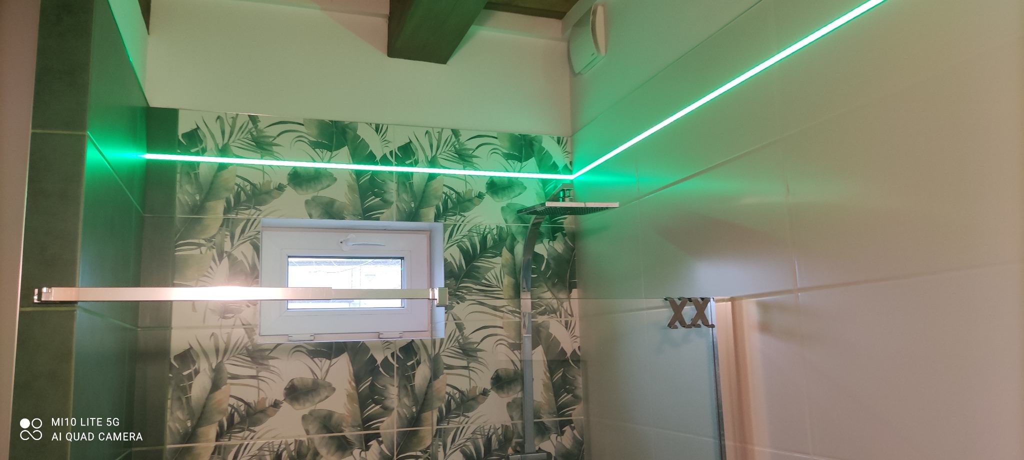 Nowoczesna łazienka z zielonym podświetleniem LED, płytkami w tropikalne liście i kabiną prysznicową typu walk-in z deszczownicą.