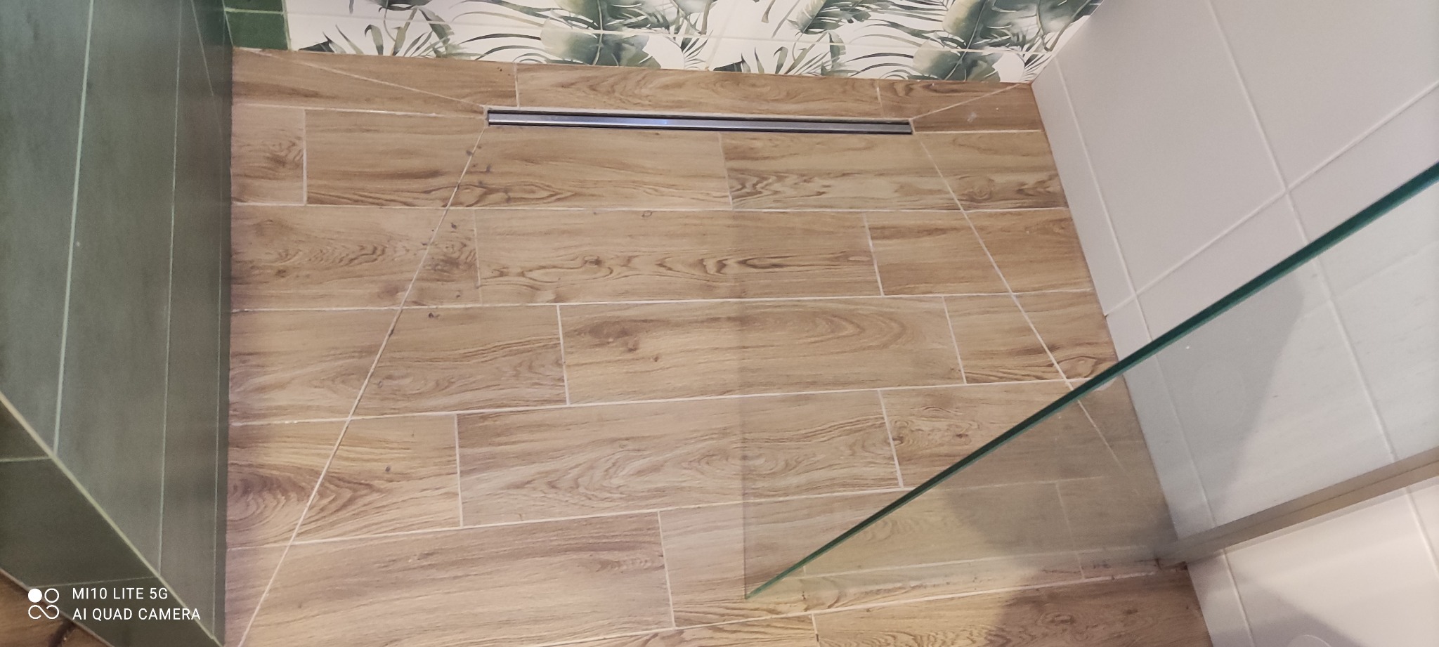 Ukończona kabina prysznicowa z odpływem liniowym i płytkami imitującymi drewno na podłodze i częściowo na ścianach, widok z góry na podłogę.