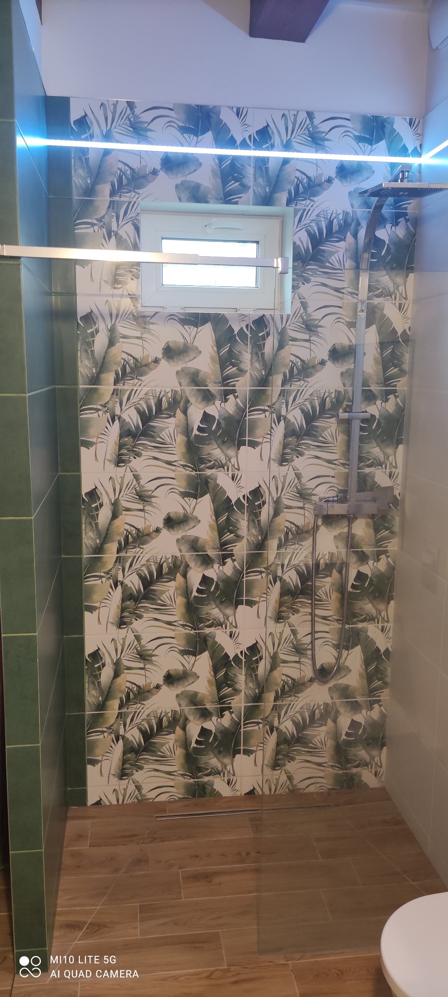 Ukończona kabina prysznicowa z deszczownicą, wyłożona płytkami z motywem tropikalnych liści, podświetlona listwą LED, z drewnopodobną podłogą i małym oknem. Widoczny fragment toalety.