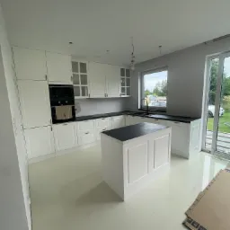 Jasna kuchnia z białymi meblami i czarnym blatem, wyspa kuchenna, widoczne niedokończone podłączenia oświetlenia, karton na podłodze.