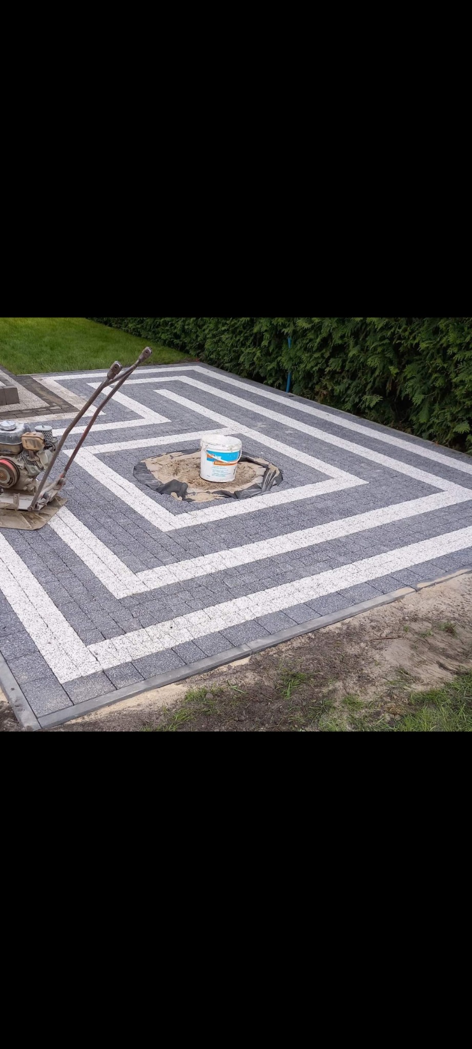 Ułożona kostka brukowa w geometryczny wzór z szarych i białych elementów, widoczna zagęszczarka w rogu i wiadro z klejem w centralnym otworze.