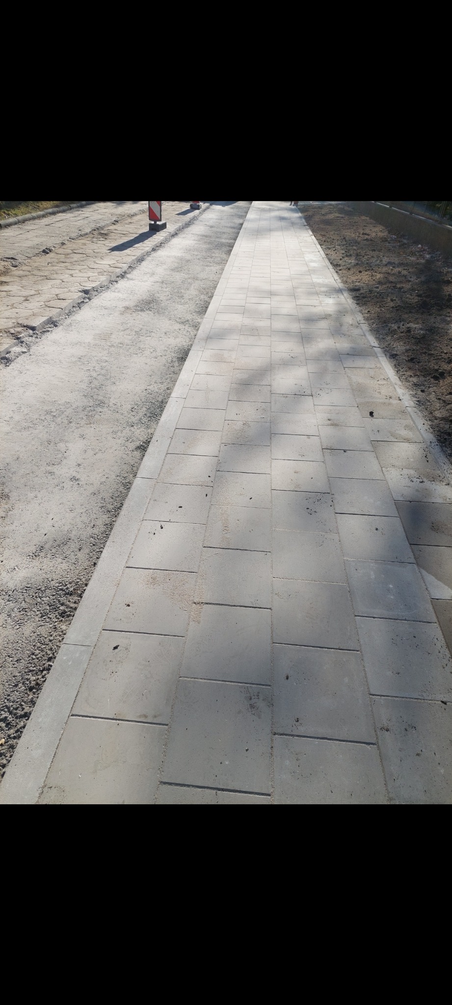 Świeżo ułożony chodnik z szarych, kwadratowych płyt betonowych, z widocznym przygotowanym podłożem po lewej stronie i fragmentem trawnika po prawej, w tle widoczne bariery drogowe.
