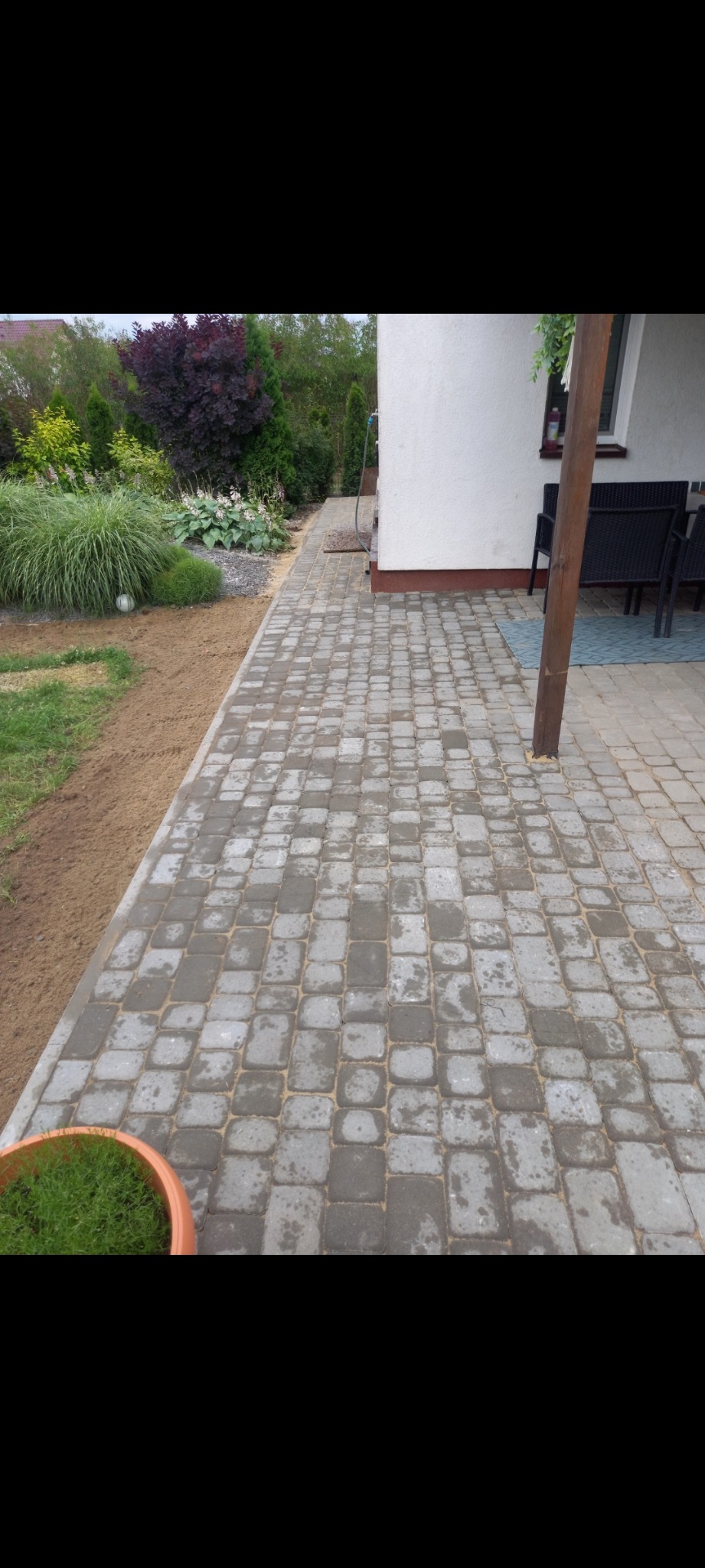 Szara kostka brukowa ułożona na posesji, widok na obrzeże z roślinnością i fragment domu z zadaszeniem.