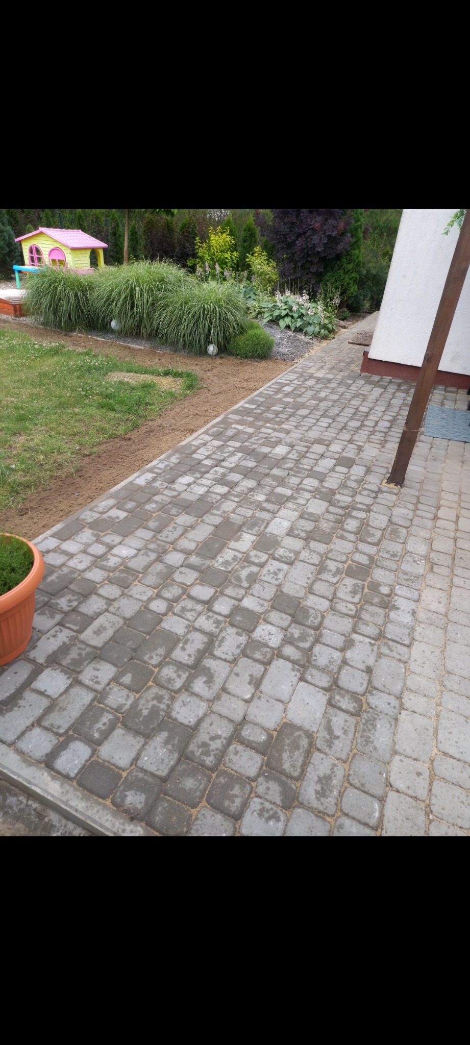 Świeżo ułożona, szara kostka brukowa na ścieżce ogrodowej, widoczne ślady wilgoci, z boku trawnik z ozdobnymi trawami i domek dla dzieci w tle.