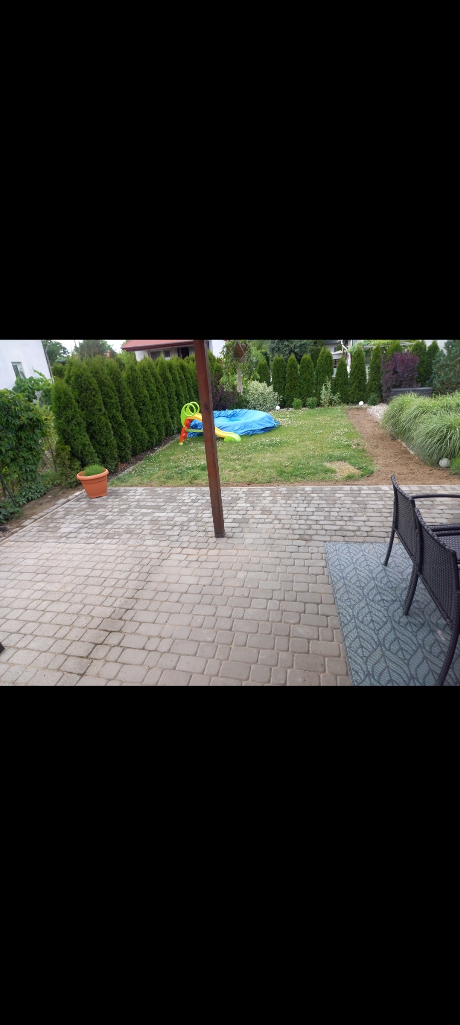 Szara kostka brukowa ułożona na posesji z widocznym trawnikiem, tujami i rozłożonym basenem dla dzieci w tle.