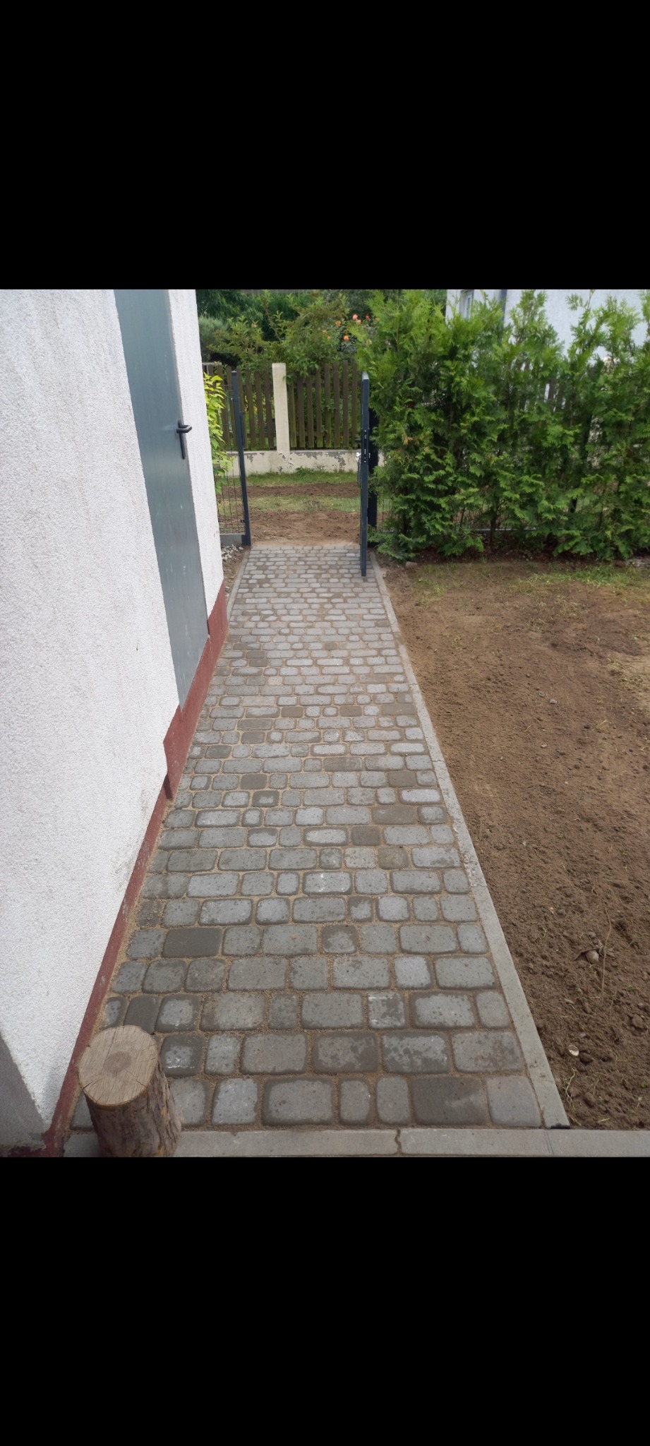 Ułożona ścieżka z szarej kostki brukowej prowadząca od ściany budynku z szarą furtką do trawnika, widoczny fragment pnia drzewa przy ścianie.