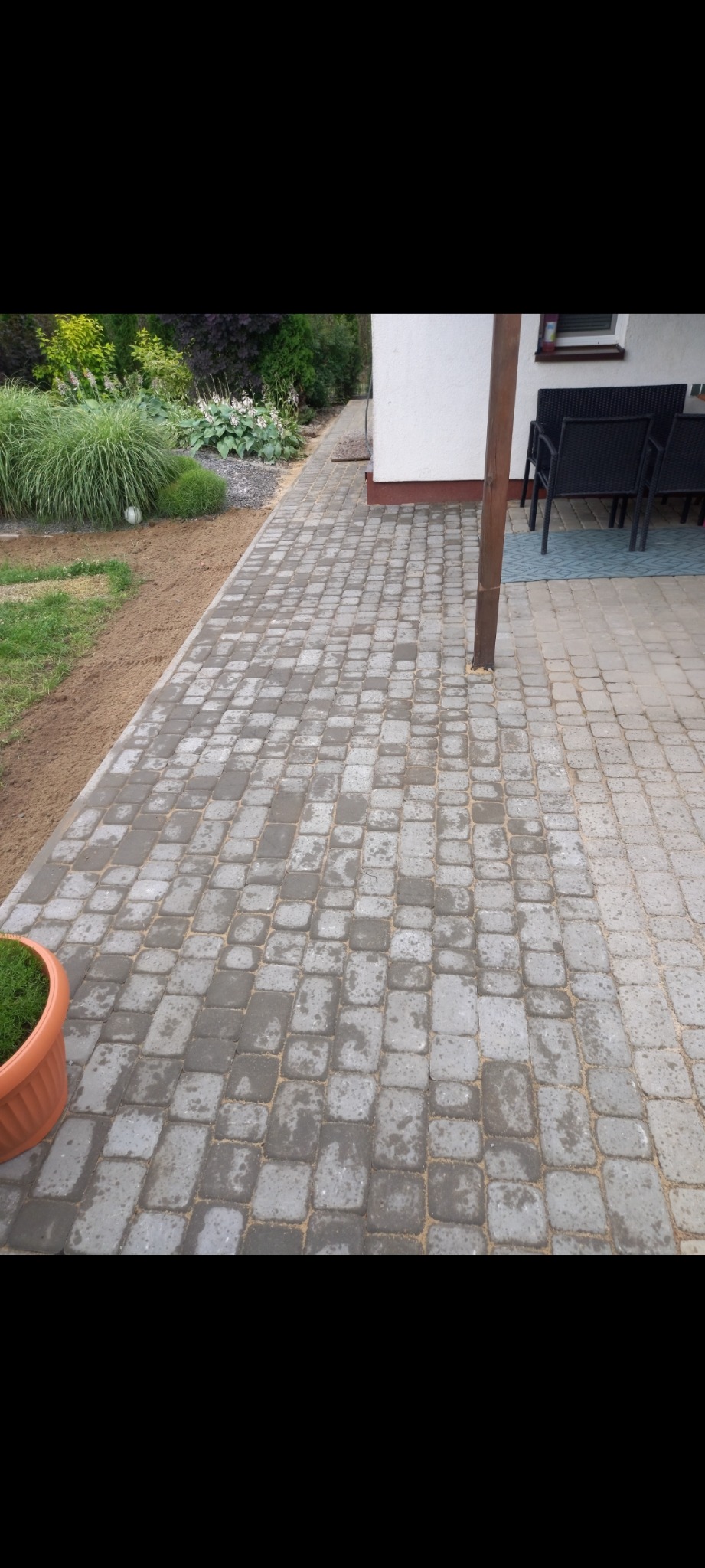 Szara kostka brukowa ułożona na posesji przylegająca do trawnika i budynku, widoczne ślady piasku w fugach, obok donica z trawą.