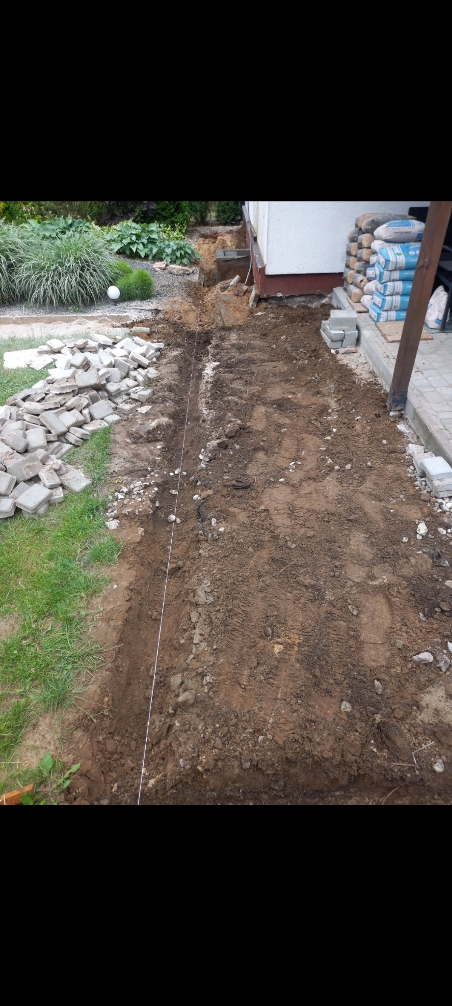 Przygotowanie terenu pod ścieżkę: wykopana ziemia, rozciągnięty sznurek wyznaczający linię, sterta kostki brukowej i worki z cementem w tle.