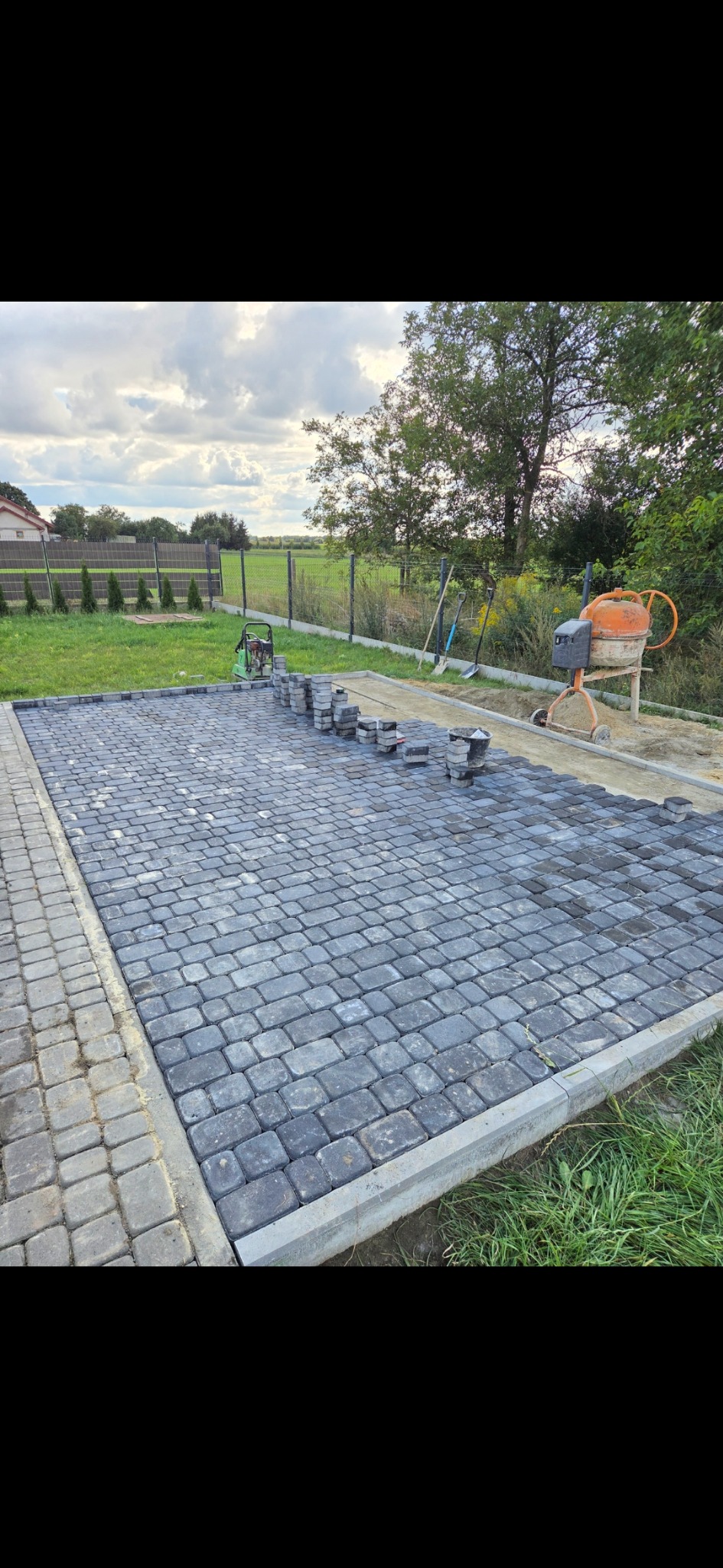 Częściowo ułożona, szara kostka brukowa na posesji. Widoczna betonowa obwódka, maszyna do mieszania betonu i narzędzia ogrodowe w tle.