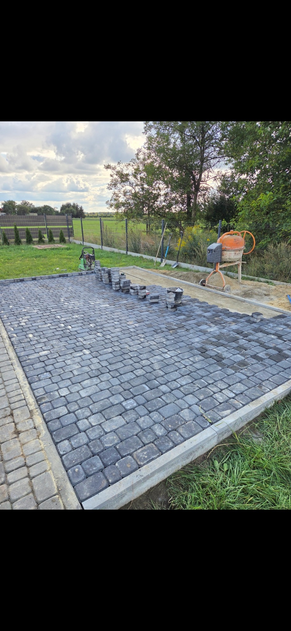 Częściowo ułożona, szara kostka brukowa na posesji. Widoczna betoniarka, ubijak, narzędzia i trawnik. Kostka obramowana szarym obrzeżem.