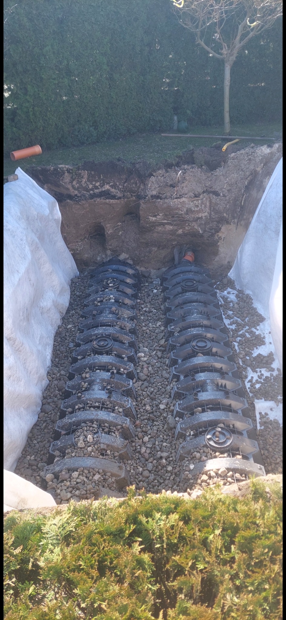 Drenaż rozsączający: Rzędy czarnych modułów w wykopie, obsypane żwirem, zabezpieczone geowłókniną. Widoczny fragment rury i zieleni w tle.