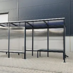 Zadaszona wiata przystankowa z siedziskiem, wykonana z metalu i przezroczystego tworzywa sztucznego, na tle szarej ściany hali magazynowej i schodów.