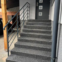 Zewnętrzne schody wejściowe wyłożone ciemnym, ziarnistym materiałem, z szarą, prostą barierką po lewej stronie, prowadzące do szarych drzwi z kwadratowymi zdobieniami.