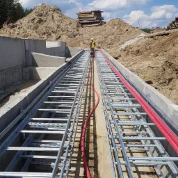 Instalacja koryta kablowego z czerwonymi przewodami w wykopie, pracownik w odblaskowej kamizelce na tle hałdy ziemi i błękitnego nieba.