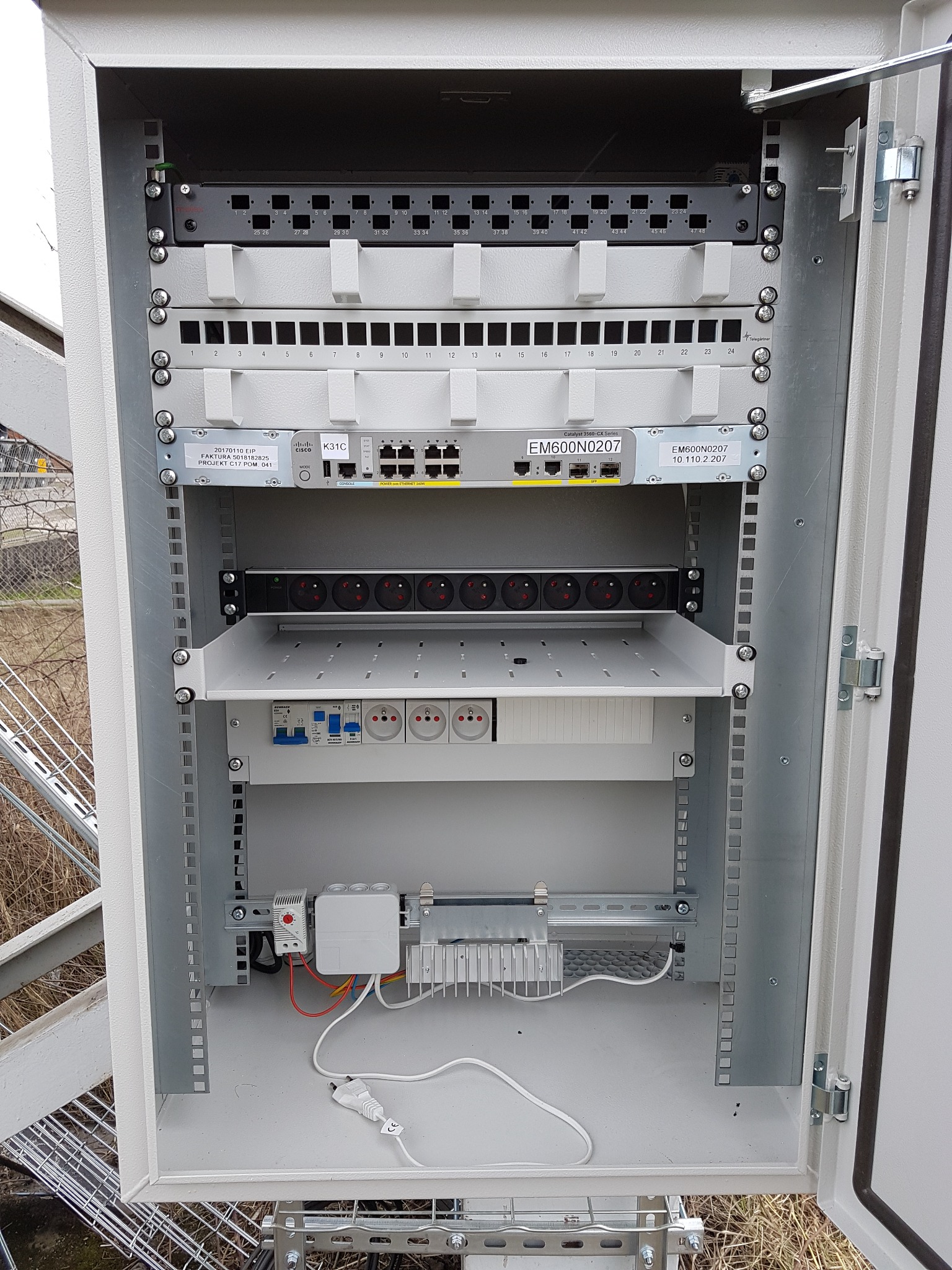 Otwarta szafa sterownicza z zamontowanymi elementami instalacji elektrycznej i sieciowej, widoczne patch panele, switch Cisco Catalyst, listwa z gniazdami, zabezpieczenia i zasilacz, na zewnątrz...