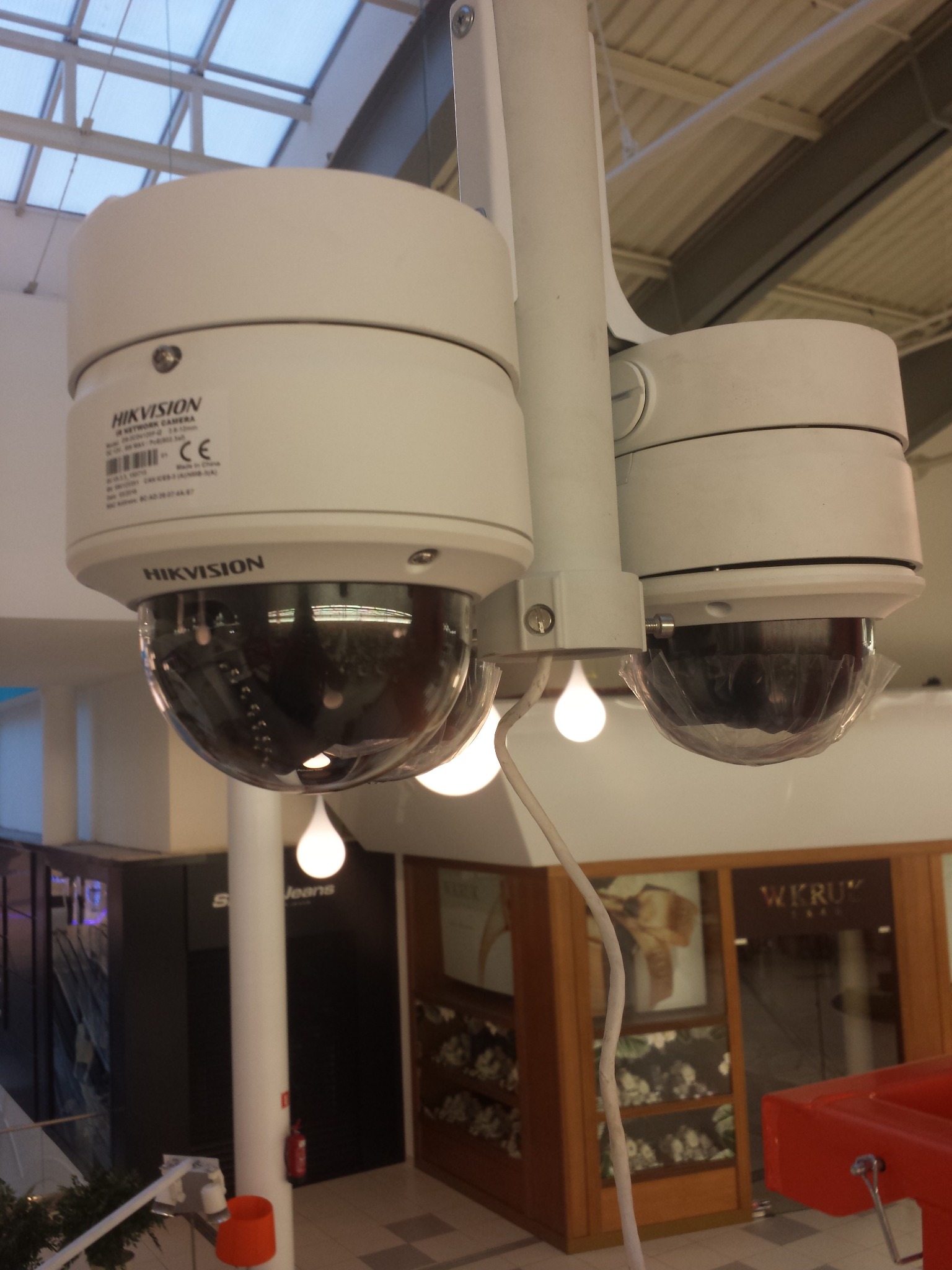 Dwie białe kamery monitoringu Hikvision, zamontowane pod szklanym sufitem w centrum handlowym z widocznymi sklepami w tle.