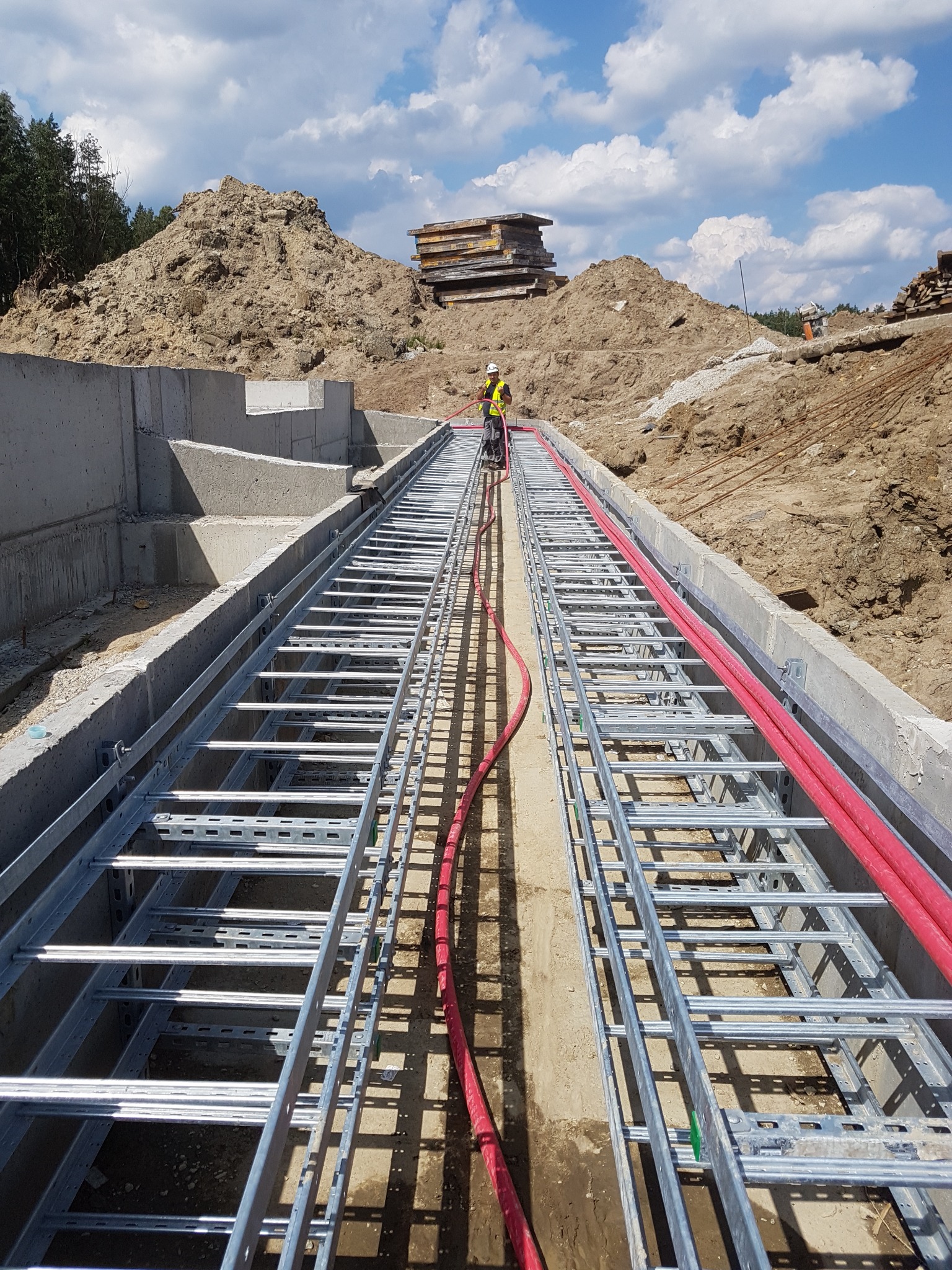 Instalacja koryta kablowego z czerwonymi przewodami w wykopie, pracownik w odblaskowej kamizelce na tle hałdy ziemi i błękitnego nieba.