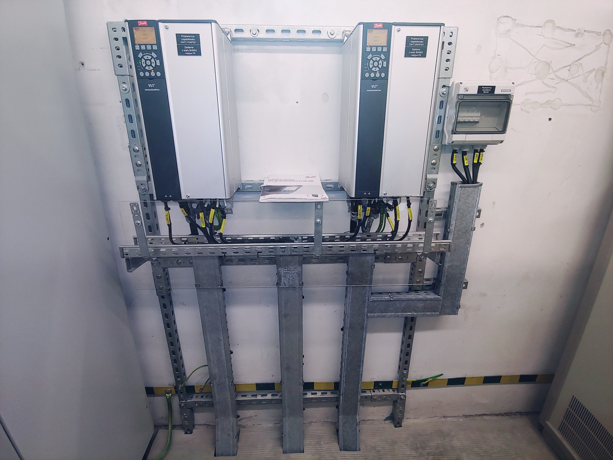 Dwa falowniki Danfoss VLT zamontowane na metalowej konstrukcji w pomieszczeniu technicznym, z widocznymi przewodami i dokumentacją techniczną, na tle ściany z widocznymi śladami po instalacjach.