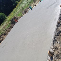 Syn-Bud - Świeżo wylana, gładka betonowa płyta fundamentowa, z widocznymi drewnianymi deskami szalunkowymi i elementami instalacji, na tle zieleni i drzew