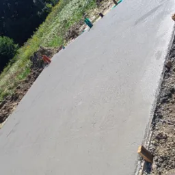 Świeżo wylana, gładka betonowa płyta fundamentowa, z widocznymi drewnianymi deskami szalunkowymi i elementami instalacji, na tle zieleni i drzew