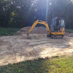 Syn-Bud - Żółta minikoparka JCB 8018 na wyrównanym, piaszczystym terenie budowy, w tle drzewa i trawa, widoczny wykop pod fundamenty i metalowa skrzynia.