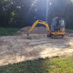 Żółta minikoparka JCB 8018 na wyrównanym, piaszczystym terenie budowy, w tle drzewa i trawa, widoczny wykop pod fundamenty i metalowa skrzynia.
