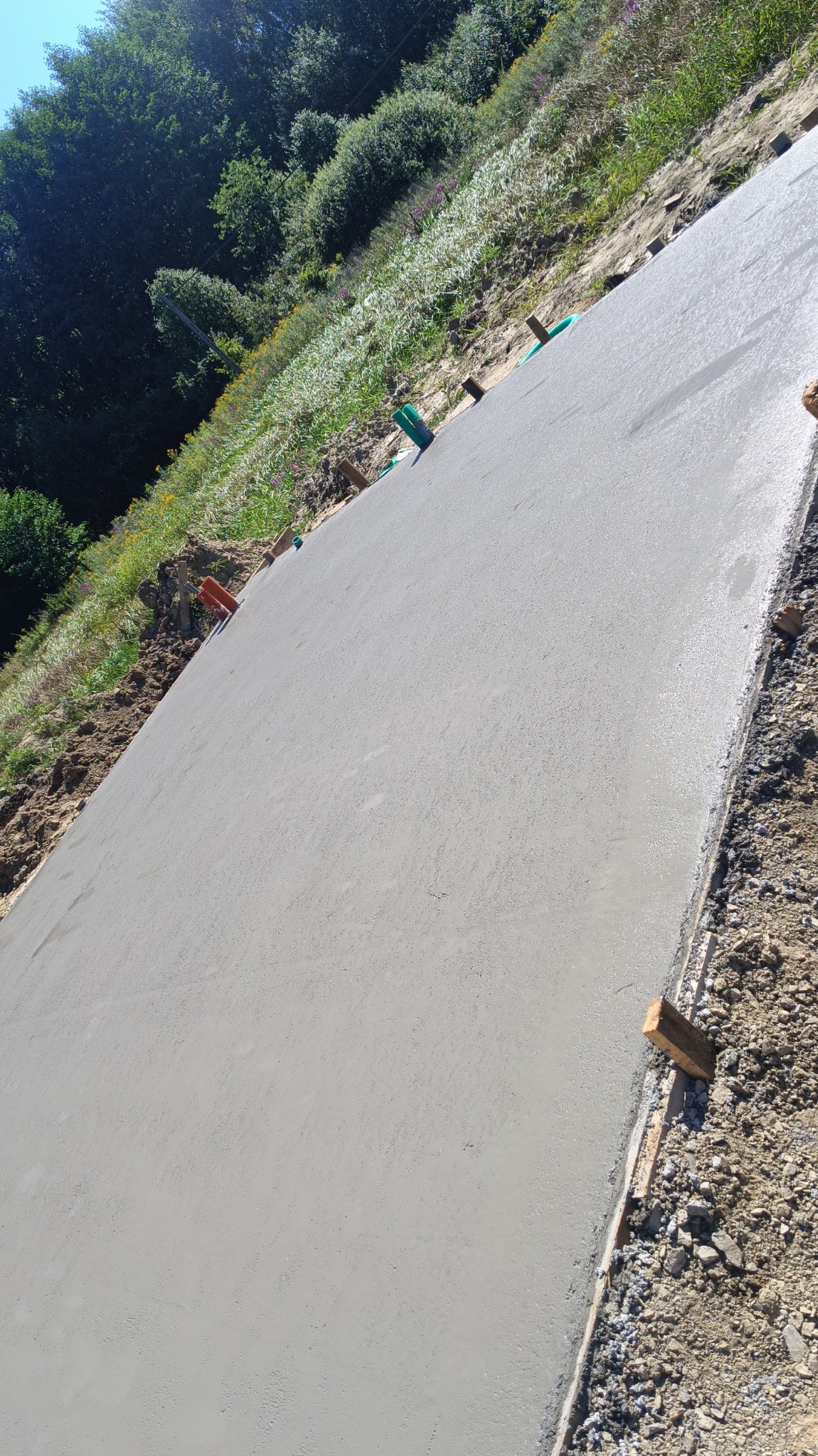 Świeżo wylana, gładka betonowa płyta fundamentowa, z widocznymi drewnianymi deskami szalunkowymi i elementami instalacji, na tle zieleni i drzew