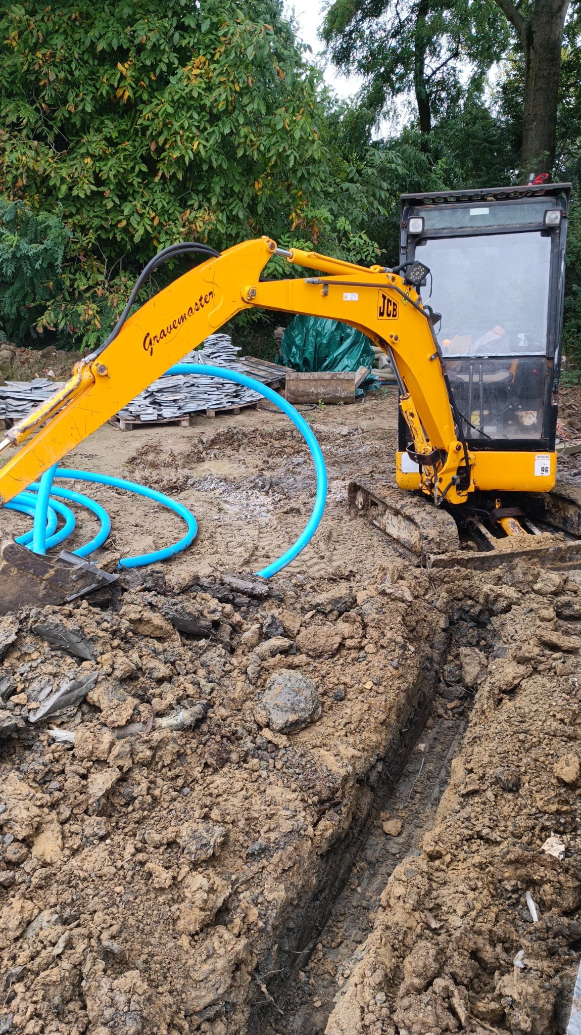 Żółta koparka JCB przy wykopanym rowie w ziemi, z niebieskimi rurami leżącymi obok, w tle widać zieloną roślinność.