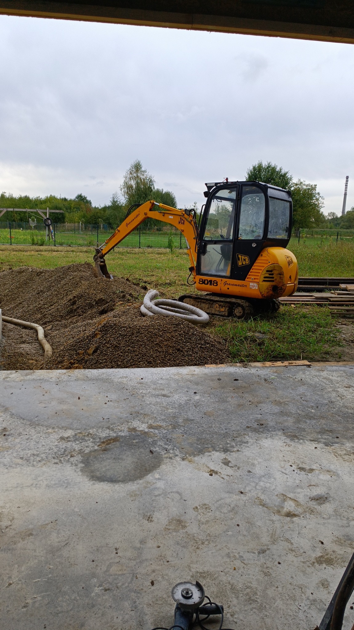 Żółta mini koparka JCB 8018 na placu budowy, obok sterta ziemi i białe rury, w tle drzewa i komin.