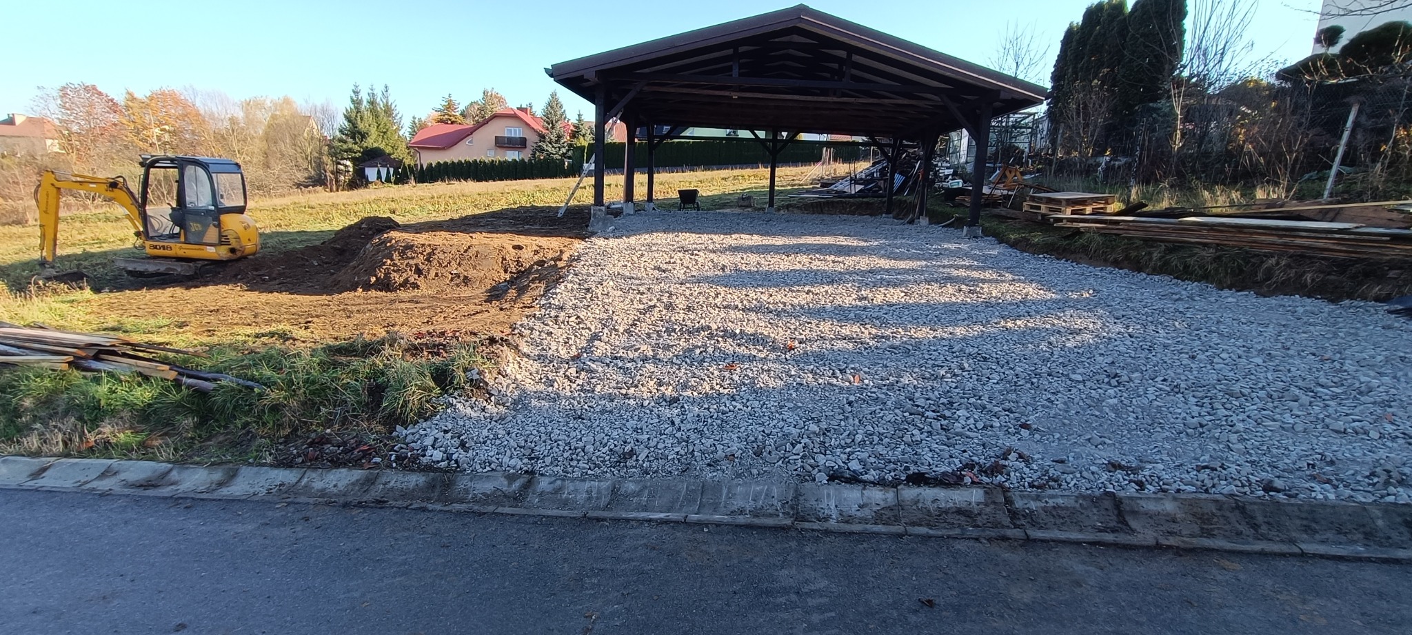 Żółta minikoparka przy pryzmie ziemi obok przygotowanego podłoża z kruszywa pod zadaszeniem, w tle dom jednorodzinny i drzewa.