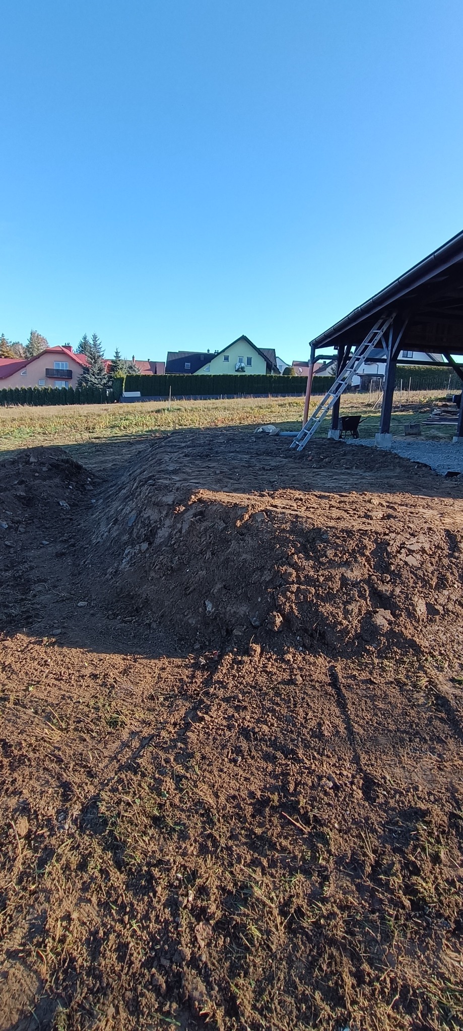 Przygotowany teren pod budowę, widoczna pryzma ziemi i wykop, w tle zabudowa jednorodzinna oraz zadaszenie z drabiną.