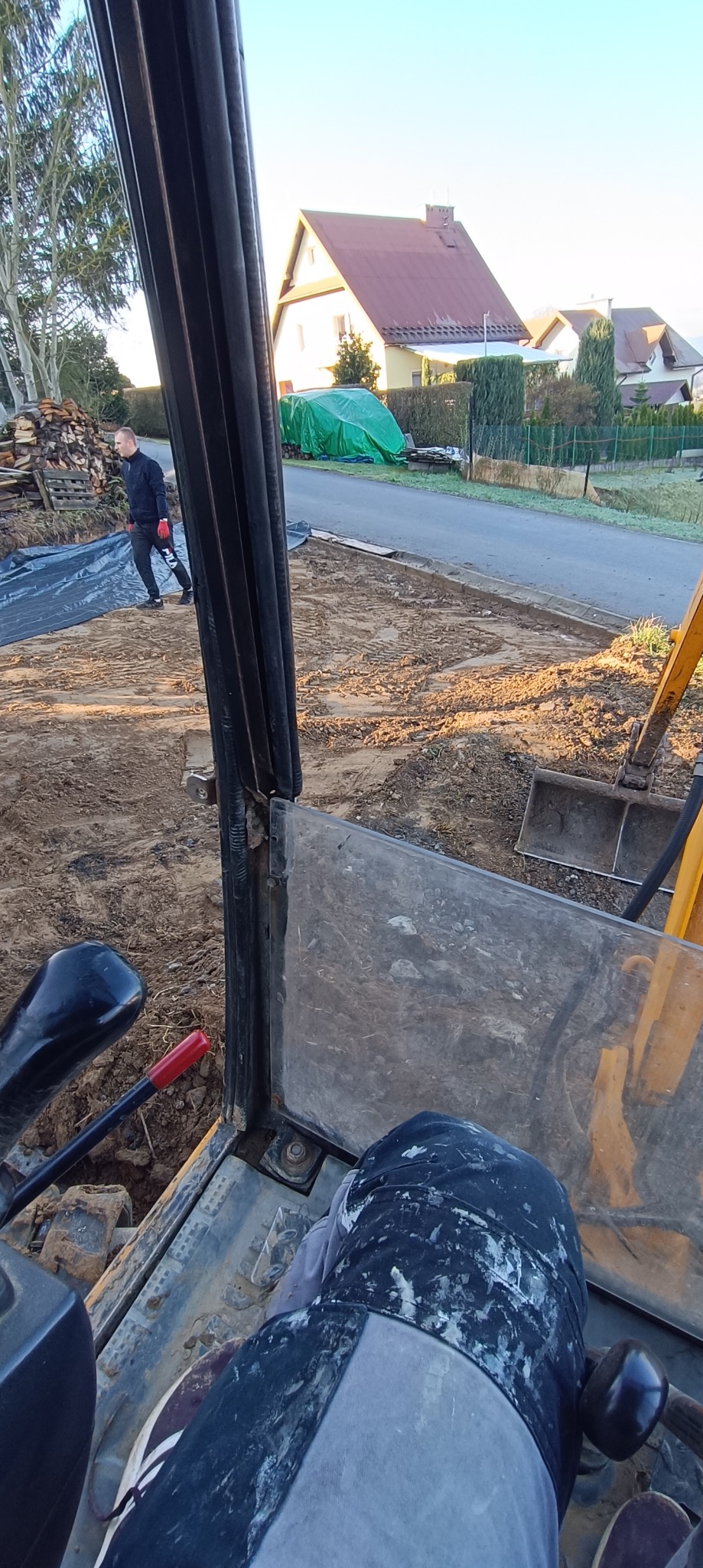 Widok z kabiny koparki na teren budowy z przygotowanym wykopem, widoczny pracownik w oddali i zabudowa jednorodzinna w tle.