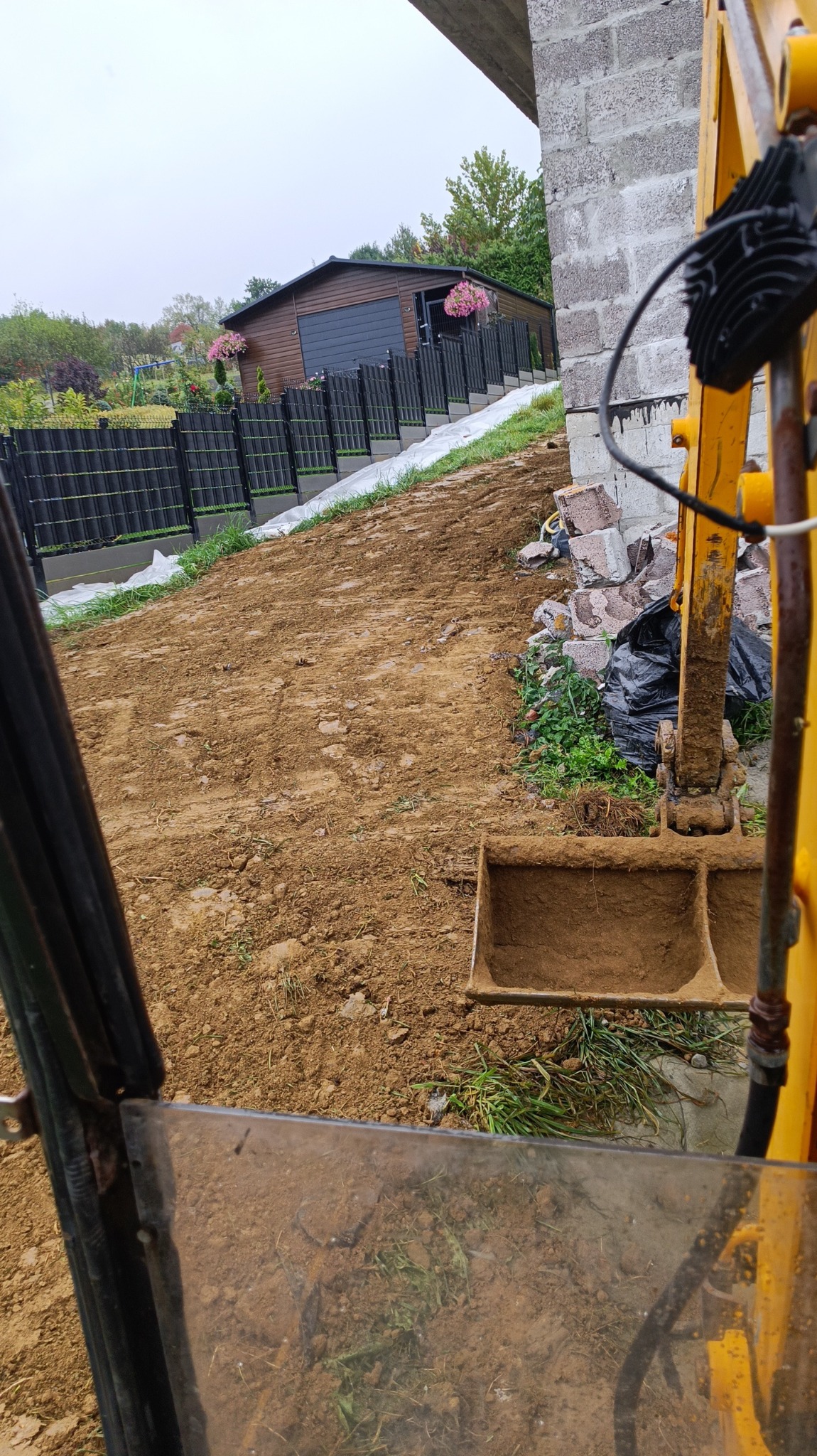 Widok z kabiny żółtej maszyny budowlanej na przygotowany teren pod budowę, z widocznym ogrodzeniem i budynkiem w tle. Częściowo wykopana ziemia z pozostałościami roślinności.