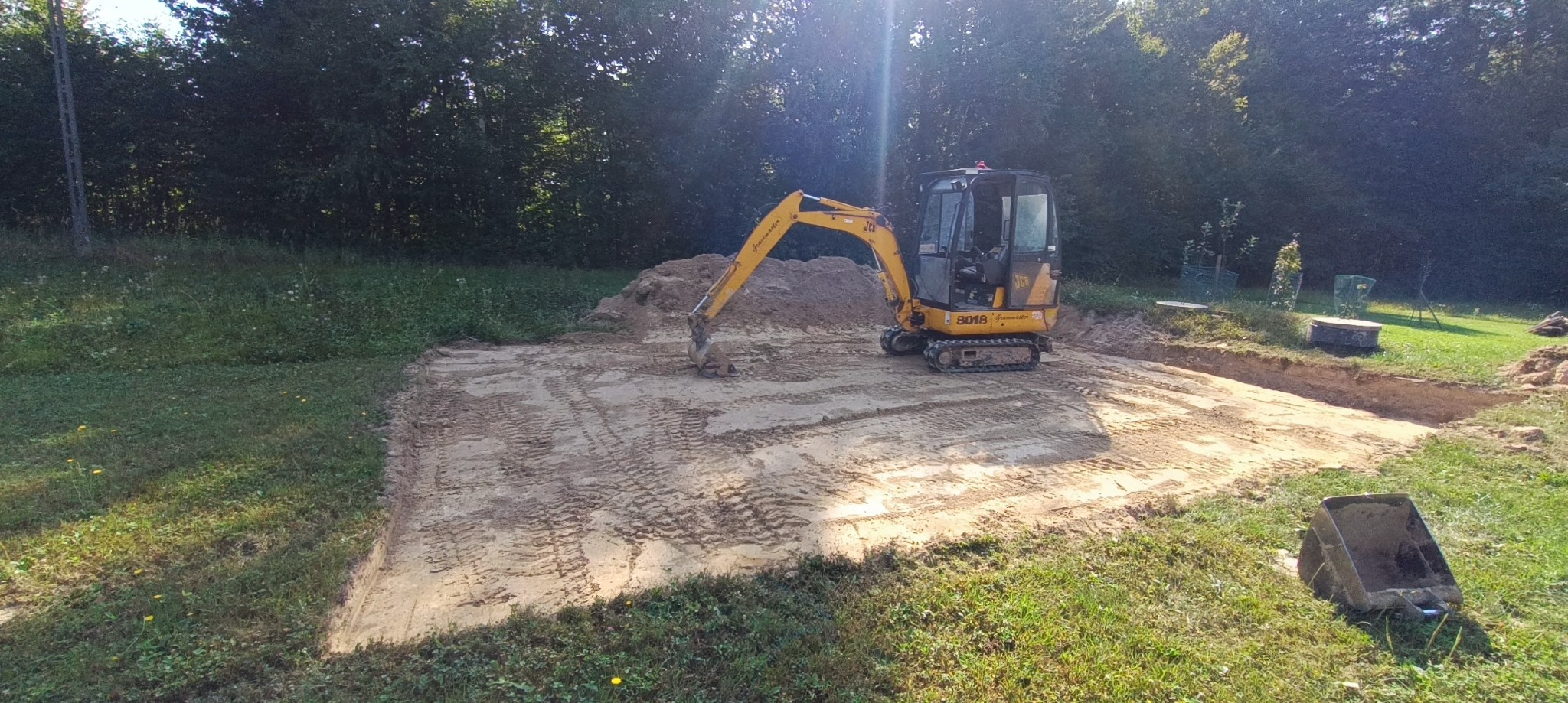 Żółta minikoparka JCB 8018 na wyrównanym, piaszczystym terenie budowy, w tle drzewa i trawa, widoczny wykop pod fundamenty i metalowa skrzynia.