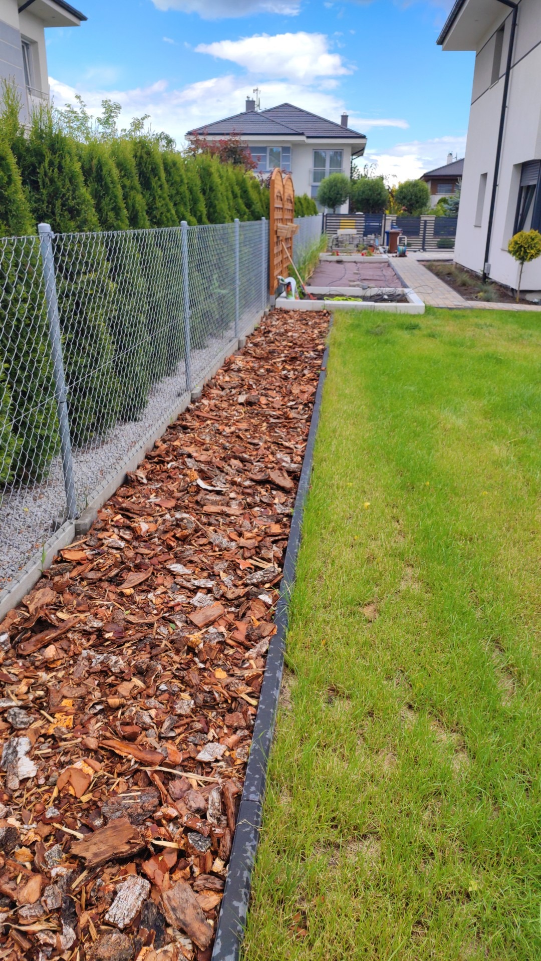 Świeżo wyłożona kora dekoracyjna wzdłuż ogrodzenia z siatki, oddzielona obrzeżem od trawnika, w tle zadbany ogród i nowoczesny dom.