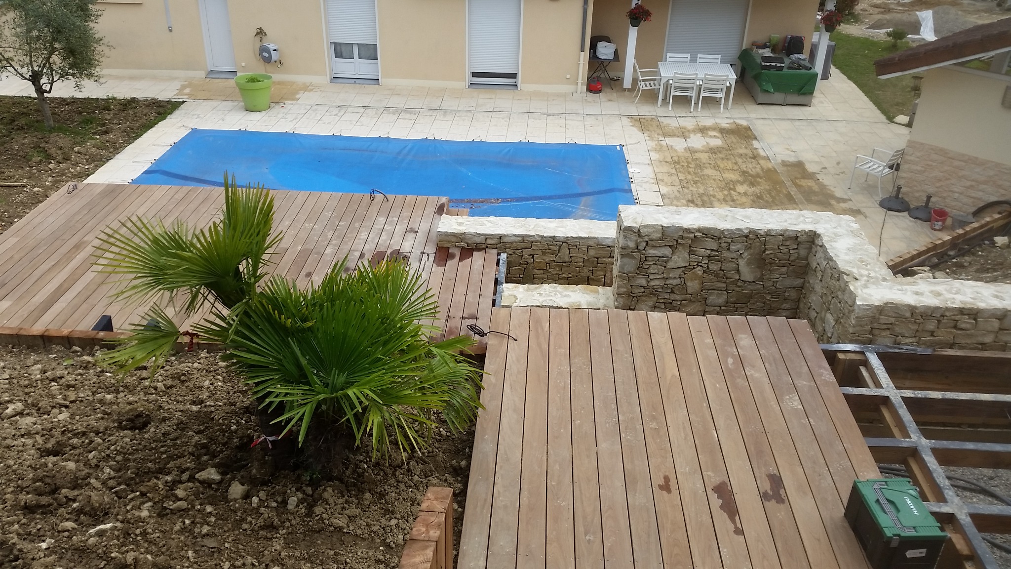 Widok z góry na taras z drewna egzotycznego w trakcie budowy, z widocznym zarysem konstrukcji nośnej, obok basen przykryty niebieską plandeką i kamienny murek, palma na pierwszym planie.