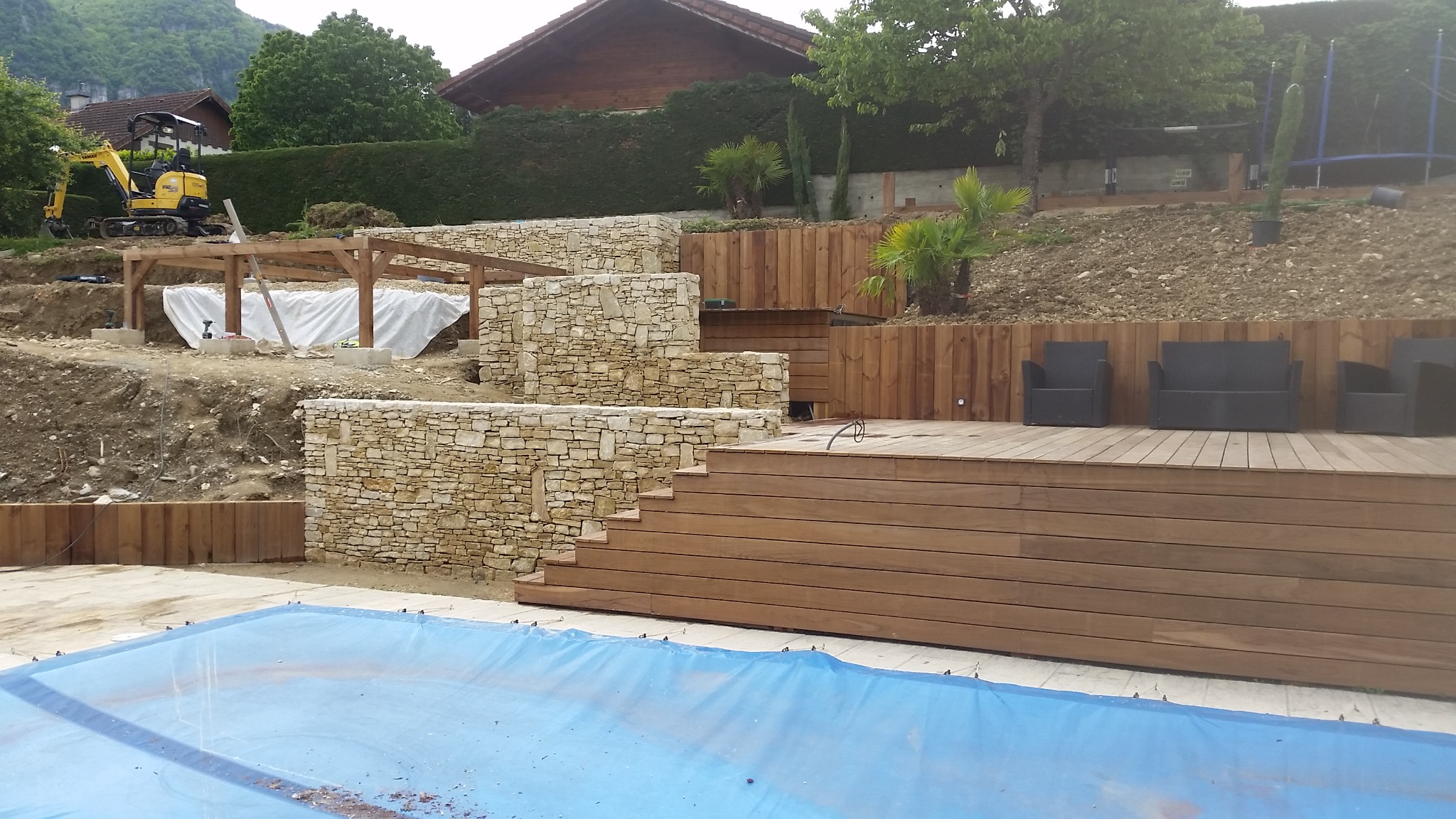 Budowa tarasowanego ogrodu z kamiennymi murkami oporowymi, drewnianą platformą z meblami ogrodowymi i basenem przykrytym plandeką, w tle koparka i częściowo zadaszona konstrukcja.