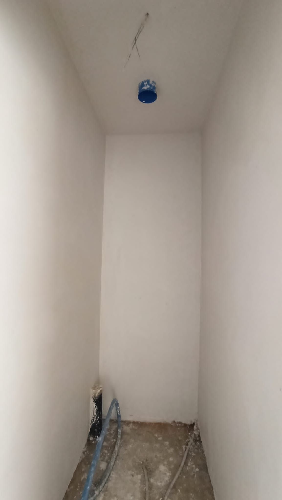 Wnętrze małego pomieszczenia po nałożeniu gładzi, widoczne przewody elektryczne wystające z sufitu oraz rury przy podłodze.