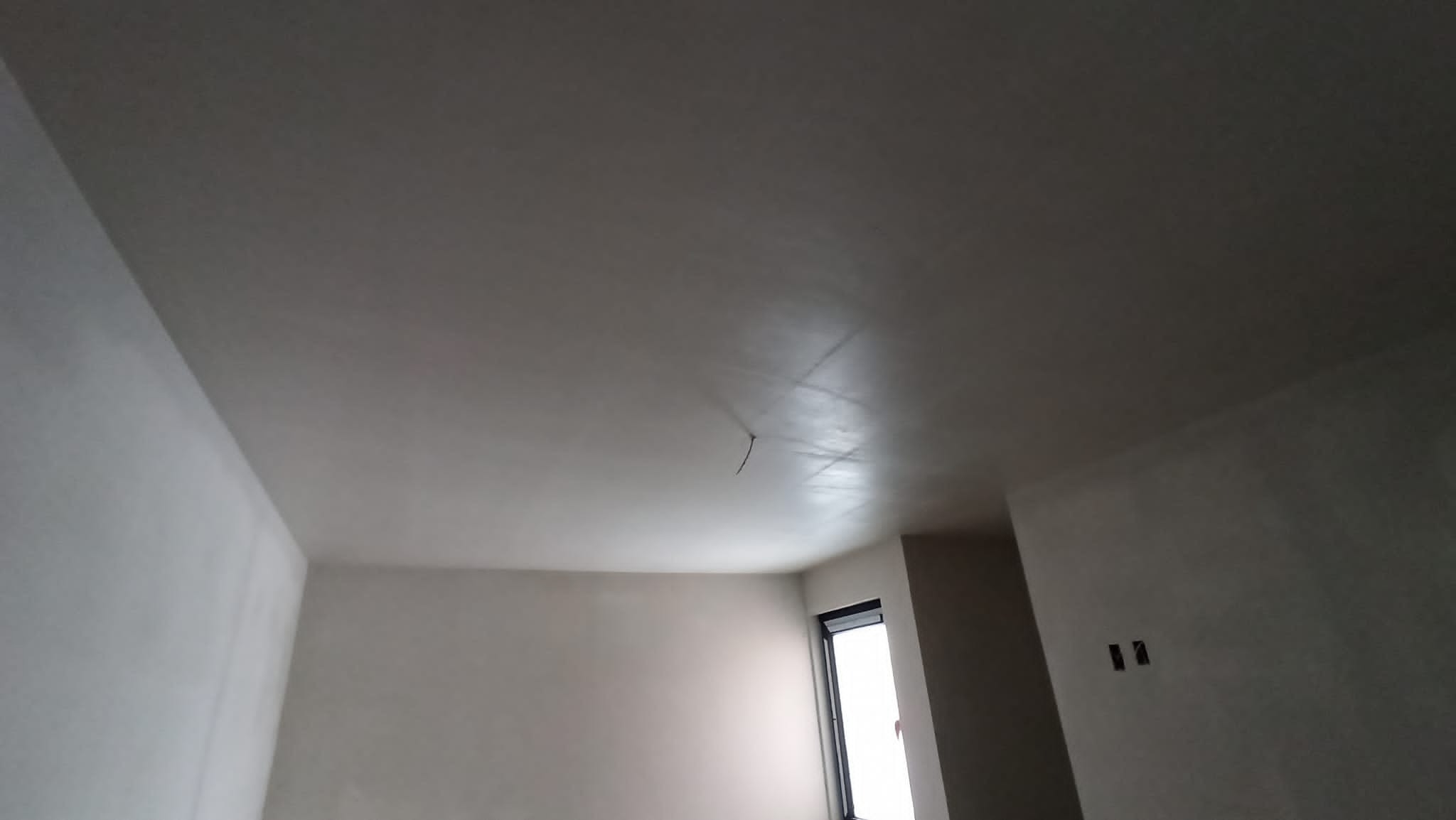 Gładko wykończony sufit po tynkowaniu maszynowym, widoczne ślady po szpachlowaniu, fragment okna z roletą i puste gniazdka elektryczne.