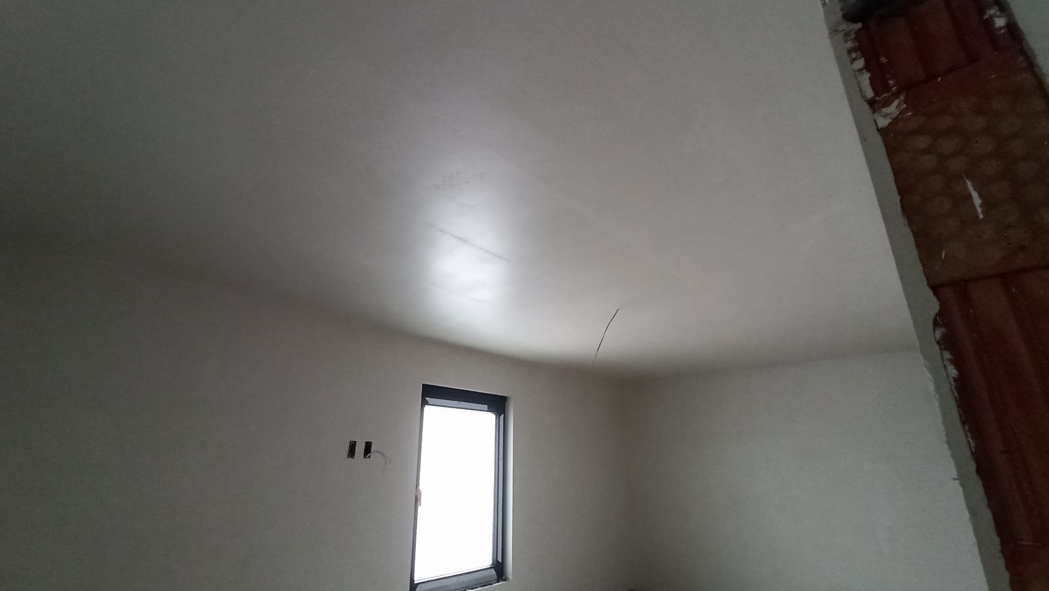 Pokój po nałożeniu gładzi, widoczne ślady po szlifowaniu na suficie, surowy stan wykończenia, okno w czarnej ramie i fragment ściany z cegły.
