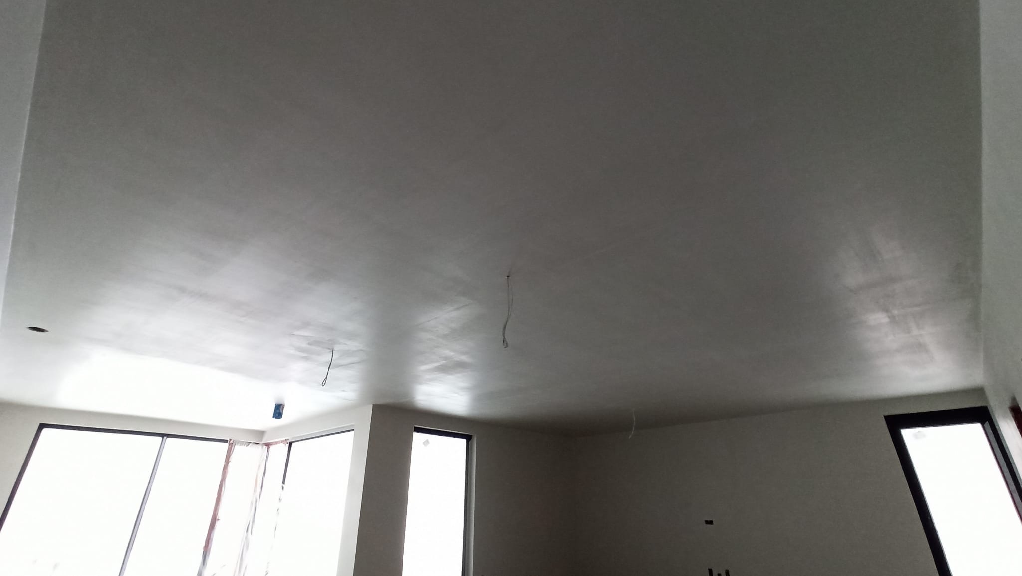 Gładka, jasnoszara powierzchnia sufitu po nałożeniu tynku maszynowego, widoczne wystające przewody elektryczne, okna bez parapetów w surowym stanie.