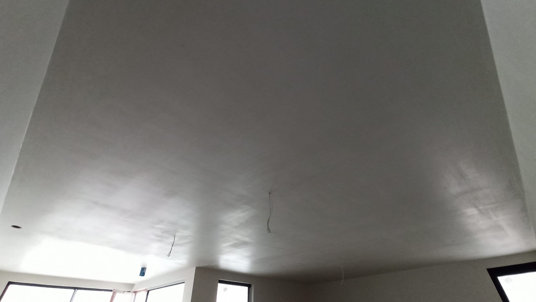 Gładka, jasnoszara powierzchnia sufitu po tynkowaniu maszynowym, widoczne przewody zwisające z sufitu i okna w tle.