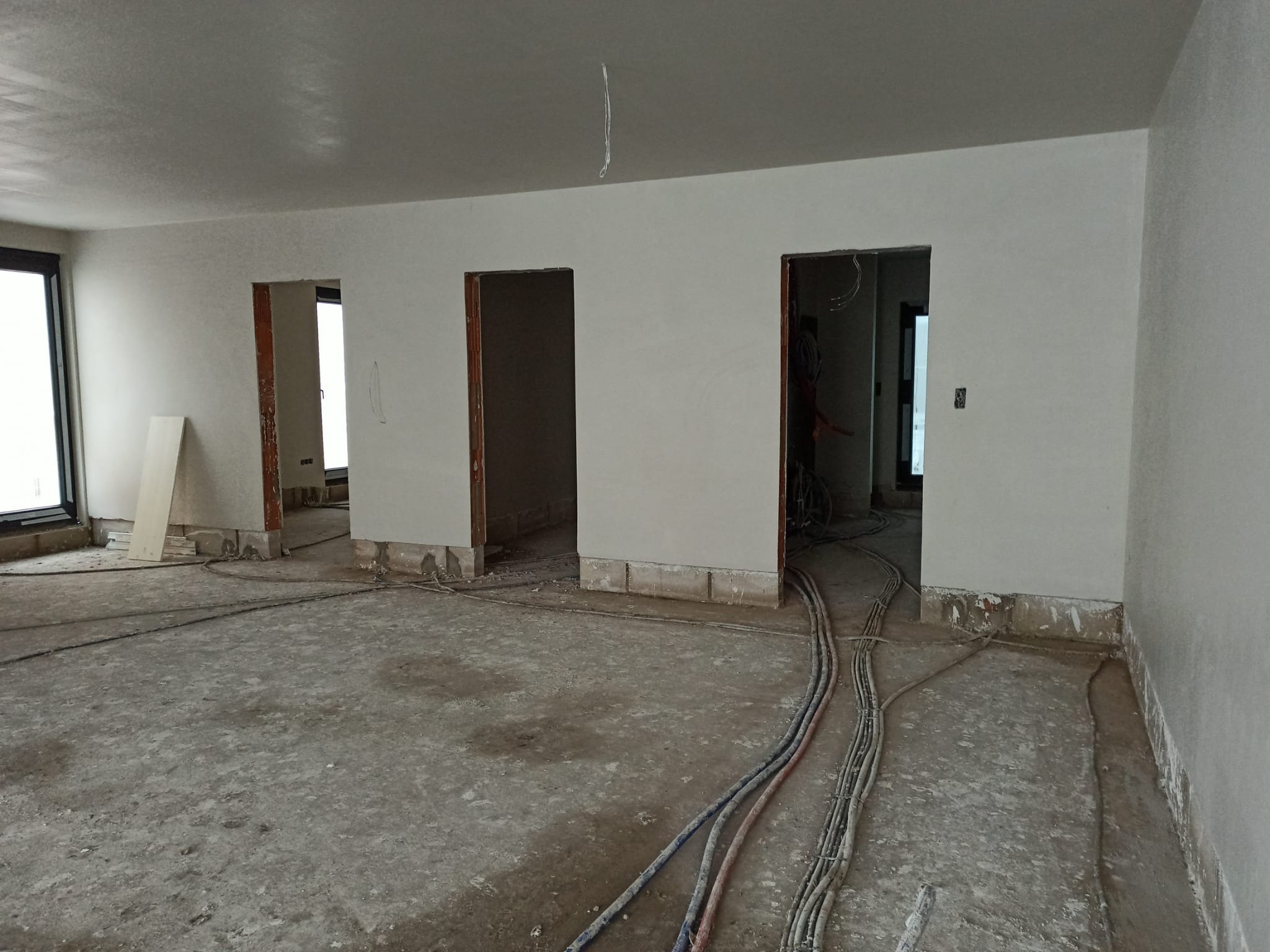 Pokój w trakcie tynkowania maszynowego: widoczne świeżo otynkowane ściany, surowa podłoga z kablami, oraz przygotowane otwory drzwiowe bez ościeżnic.