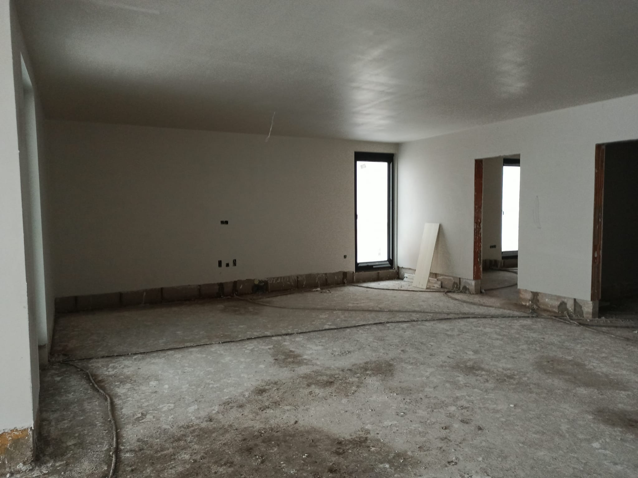 Wnętrze w trakcie prac tynkarskich: świeżo otynkowane ściany i sufit, widoczne przewody elektryczne na podłodze, przygotowanie do dalszych prac wykończeniowych, okno z czarną ramą.