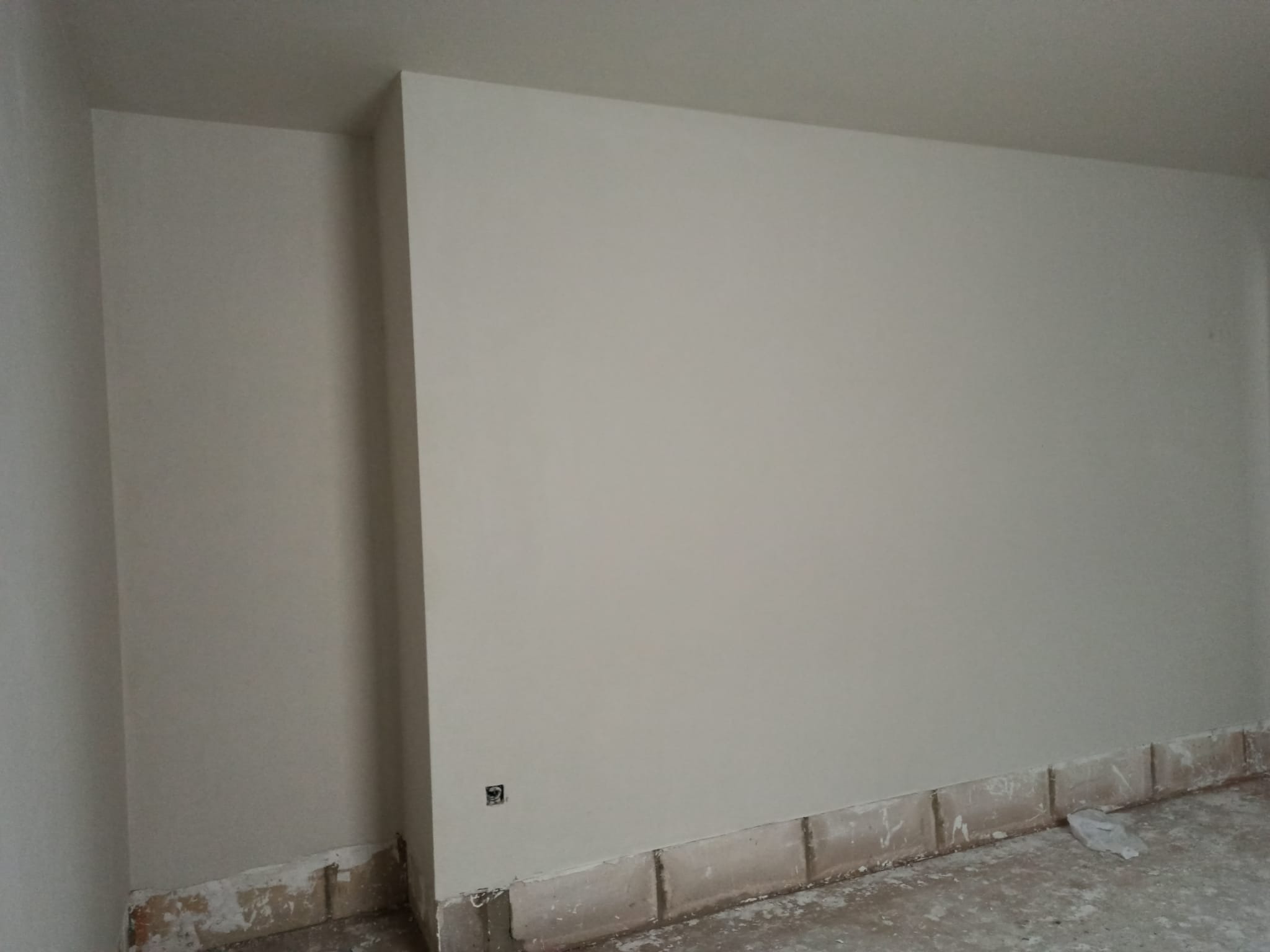 Gładko otynkowana ściana w jasnym odcieniu szarości, z widocznym gniazdkiem elektrycznym i fragmentami uszkodzonej podłogi przy ścianie, sugerującymi prace wykończeniowe.