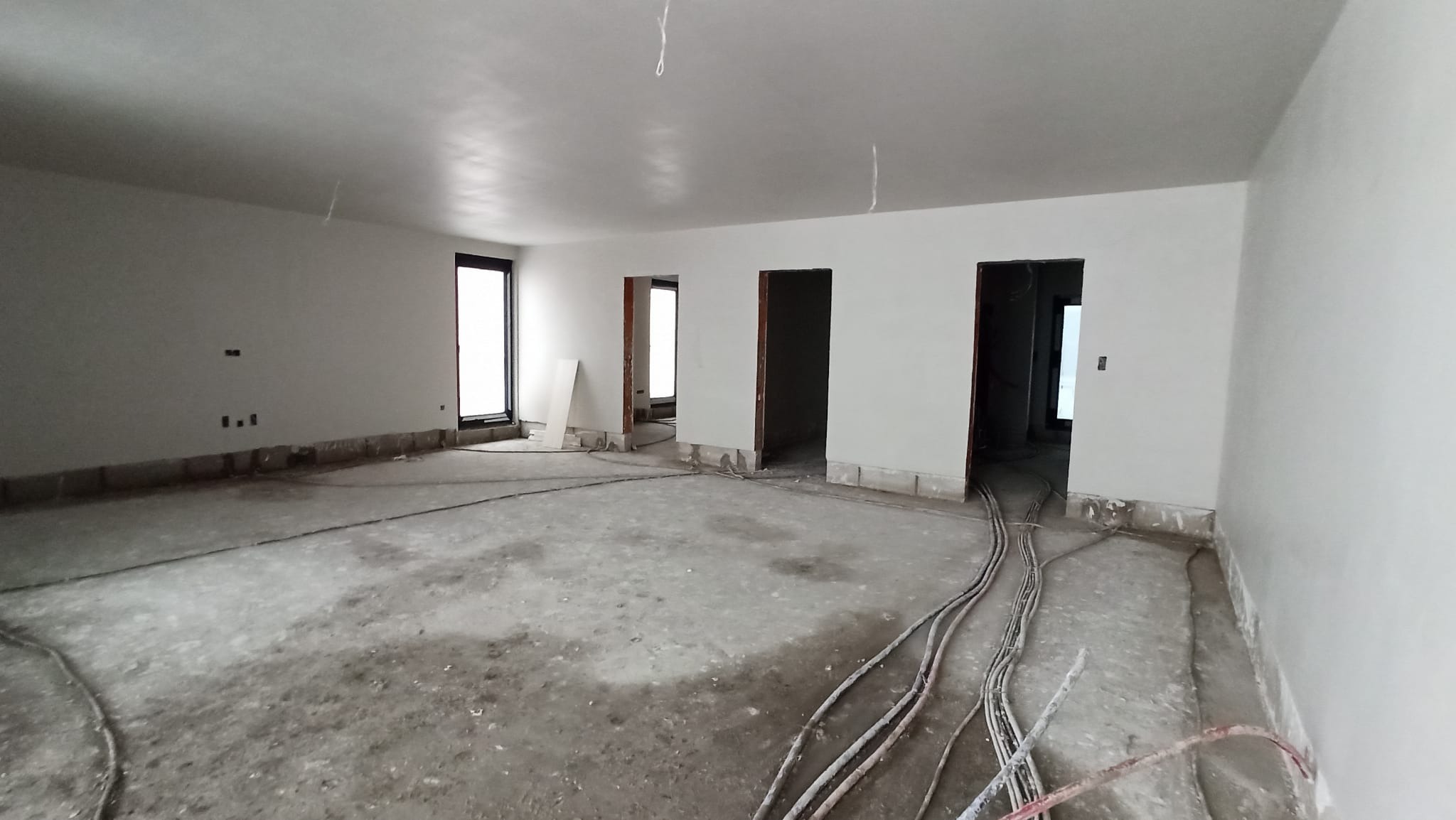 Wnętrze w stanie deweloperskim po nałożeniu gładzi gipsowej na ścianach i suficie, widoczne wystające przewody elektryczne i przygotowane otwory drzwiowe.