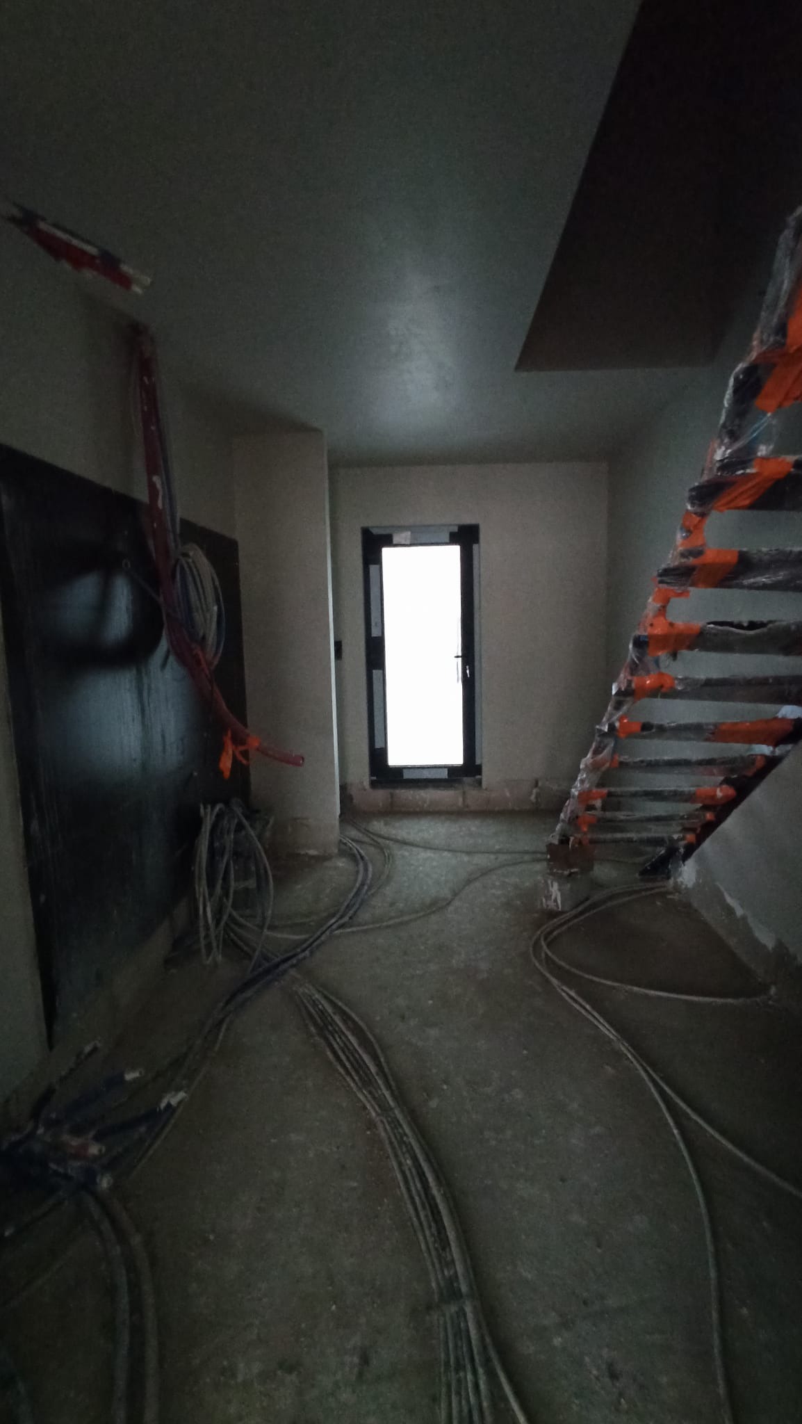 Wnętrze w stanie surowym: przewody elektryczne na podłodze, ściany przygotowane do wykończenia, schody zabezpieczone folią, okno w czarnej ramie w tle.